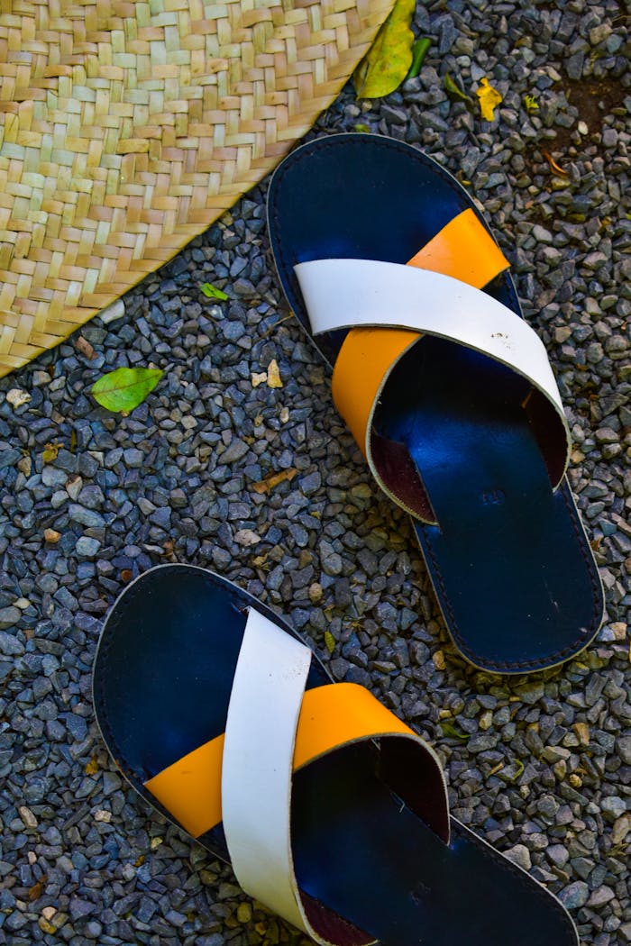 Pair of colorful sandals on a pebbled ground next to a straw hat.